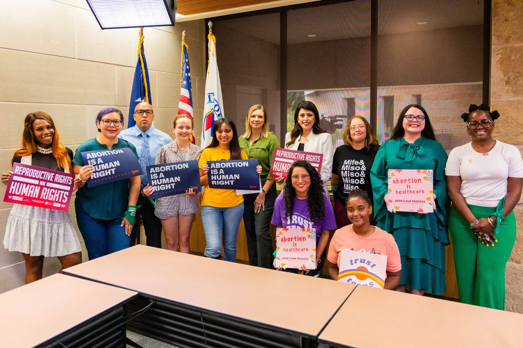 Ipas and partners with Austin City Councilwoman, Vanessa Fuentes A group of eleven people standing together indoors, holding various signs supporting abortion rights. Some signs read "Abortion is a Human Right" and "Reproductive Rights." They are in front of flags and smiling for the photo.